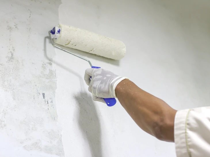 Peintre professionnel en combinaison bleue et casquette blanche applique de la peinture blanche au rouleau sur un mur intérieur