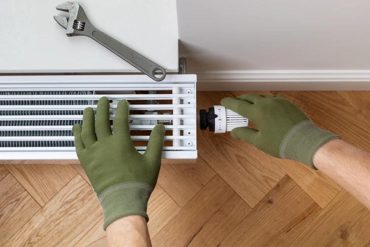 Intérieur résidentiel avec technicien en combinaison rouge et bleue installant un radiateur blanc à ailettes sur mur gris