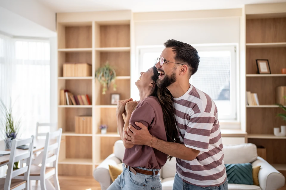 Scène d'intérieur professionnel avec deux personnes en costume échangeant une maquette de maison miniature au-dessus de documents contractuels